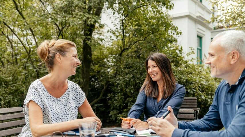 Fotografie einer Unterhaltung von zwei lachenden Frauen und eines Mannes im Garten am Tisch mit Unterlagen.