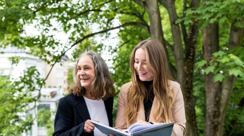 Zwei Frauen stehen draußen vor grünem Hintergrund und lachen.
